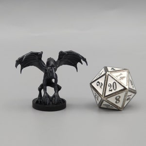 Puede incluir: Una figura en miniatura negra de una gárgola con las alas extendidas, de pie sobre una base negra. La figura es aproximadamente del tamaño de un dado de 20 caras, que también se muestra en la imagen.