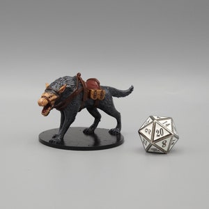 May include: A black and gray miniature wolf figurine with a brown saddle and saddlebags. The wolf is standing on a black base and is facing to the left. A silver 20-sided die is in the background.