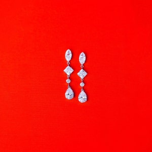 May include: A pair of silver dangle earrings with clear cubic zirconia stones. The earrings feature a pear-shaped stone at the bottom, a square stone in the middle, and a marquise-shaped stone at the top.