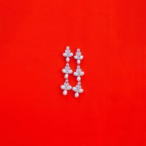 May include: A pair of silver dangle earrings with white pearl accents. The earrings have a floral design and are on a red background.