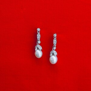 May include: A pair of silver earrings with pearl drops and clear crystal accents on a red background.