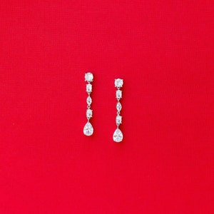 May include: A pair of silver dangle earrings with clear pear-shaped and round cubic zirconia stones on a red background.