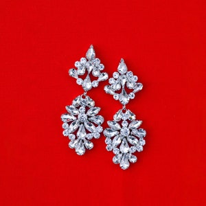 May include: A pair of silver dangle earrings with large, clear crystal stones arranged in a floral pattern. The earrings are on a red background.