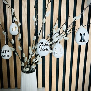 Puede incluir: Huevos de Pascua de cerámica blanca con letras doradas colgando de ramas de sauce en un jarrón blanco. Los huevos tienen el texto "Happy Easter", "Frohe Ostern" y "Hello Spring".