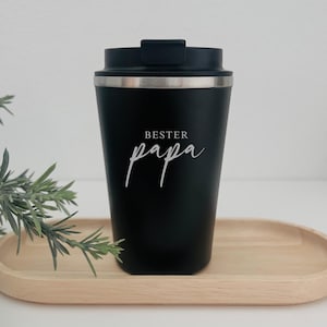 Könnte beinhalten: Ein schwarzer Reisebecher mit silbernem Rand und schwarzem Deckel. Die Worte "BESTER papa" sind in weißer Schrift auf der Vorderseite des Bechers geschrieben. Der Becher steht auf einem hellbraunen Holztablett.