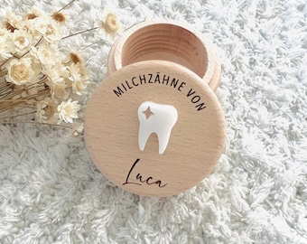 May include: A round, wooden keepsake box with a lid. The lid features the text "MILCHZÄHNE VON" and a white tooth design with a star. The name "Luca" is written below the tooth. The box is open, revealing the interior.