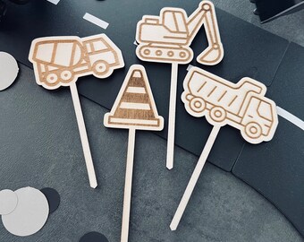 May include: Set of five wooden cake toppers featuring construction vehicles and a traffic cone. The toppers are in a line on a black surface with white and black confetti.