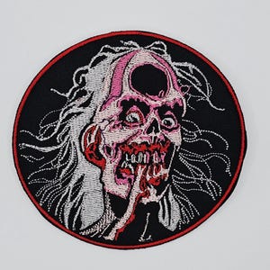 May include: Embroidered patch featuring a grotesque zombie face with exposed skull, red mouth, and pink accents. The patch is circular with a black background and a red border. The zombie has white hair and is sticking out its tongue.