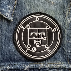 Peut inclure: Un écusson brodé noir et blanc avec un cercle contenant un symbole et les lettres "B", "O", "E", "S", "I", "L", "T", et "C". L'écusson est cousu sur un gilet en jean bleu.