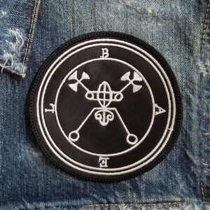 Könnte beinhalten: Ein gestickter Patch in Schwarz und Weiß mit einem Kreisdesign und den Buchstaben "B", "A", "L" und "E" um den Kreis herum. Das Design innerhalb des Kreises beinhaltet ein Symbol mit einem Kreis und zwei Sätzen gekreuzter Pfeile.