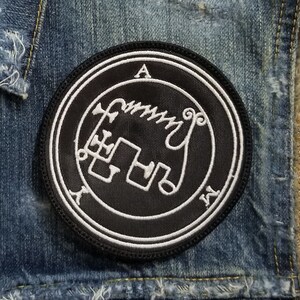 May include: A black denim vest with a white embroidered patch featuring a sigil and the letter "A".