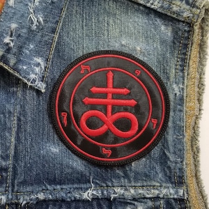 May include: A denim vest with a black and red patch featuring a symbol with a cross and an infinity sign. The vest has a distressed look with frayed edges and faded denim.