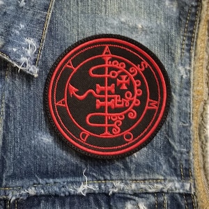 May include: A black denim vest with a red embroidered patch featuring a sigil with a serpent and the letters 'A', 'M', 'D', 'Y', and 'O'.