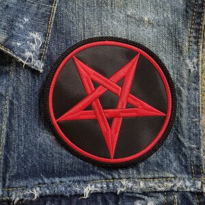 Op de afbeelding: Een denim vest met een zwarte en rode pentagram patch. De patch is genaaid op de voorkant van het vest, dichtbij de kraag.