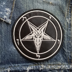 Può includere: Una toppa ricamata in bianco e nero di un pentagramma con una testa di capra all'interno. Il pentagramma è circondato da un cerchio con la lettera ebraica 'K' su ogni lato. La toppa è cucita su una giacca di jeans.