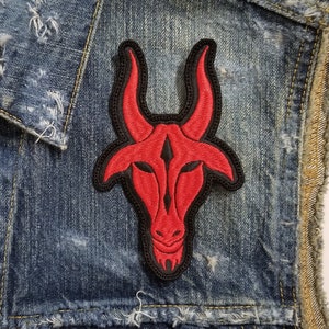 Op de afbeelding: Een rood geborduurd patch van een gehoornde stierenkop op een spijkerjack. De patch is zwart omrand en heeft een rood en zwart ontwerp.