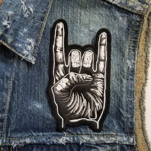 May include: A black and white iron-on patch of a hand making the rock and roll hand gesture. The patch is on a blue denim vest.