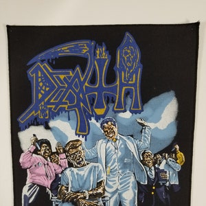 May include: A black and white patch featuring the band name "DEATH" in blue and gold lettering. The image depicts a band member in a white suit and a patient in a hospital bed. The text "Spiritual Healing" is in gold lettering. The patch is from 1990 and distributed by Razormatz.