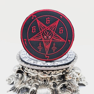 May include: A red embroidered pentagram with a goat's head inside, surrounded by the number 666, on a black circular patch. The patch is displayed on a silver decorative stand.