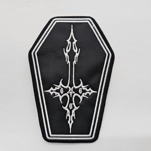 May include: Black and white embroidered patch shaped like a coffin with a white cross inside. The cross has a star at the center and the number 6 on each side.