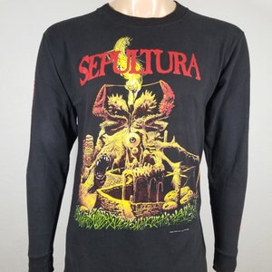 Könnte beinhalten: Schwarzes Langarmshirt mit einem gelben und roten Grafikdruck eines Schädels und einer brennenden Kerze. Der Text "SEPULTURA" ist in Rot über dem Shirt gedruckt.