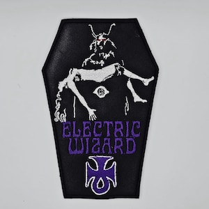 May include: Black, coffin-shaped patch with a white illustration of a horned figure and a person. The purple text reads "ELECTRIC WIZARD", with a purple cross symbol below. This patch is designed to be sewn onto clothing or bags.