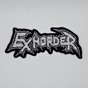 May include: A black and white embroidered patch featuring the word "EXHORDER" in a jagged, stylized font. The white text is stitched onto a black, irregularly shaped background. The design has a heavy metal theme, with a distinctive, edgy appearance.