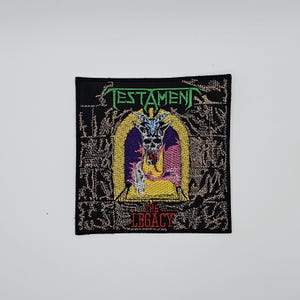 Könnte beinhalten: Schwarzer, gestickter Aufnäher mit dem Bandnamen "TESTAMENT" in Grün über einer Illustration eines gehörnten Schädels in einem gelben Torbogen. Der Text "THE LEGACY" befindet sich unter der Illustration. Der Aufnäher hat einen strukturierten Hintergrund.