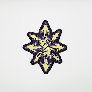 May include: Embroidered patch featuring a yellow eight-pointed star design with arrows pointing outwards, set against a black background with a purple border. The patch has a detailed, textured appearance.