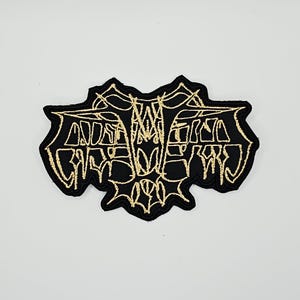 May include: A black embroidered patch featuring a stylized, symmetrical bat design outlined in metallic gold thread. The bat is depicted with wings spread wide, showcasing intricate, jagged lines and sharp points throughout the entire graphic.