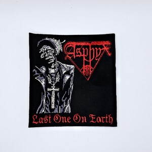 Puede incluir: Parche bordado negro con una ilustración detallada de una figura con un colgante de cruz y la palabra "Asphyx" en rojo. El texto "Last One On Earth" está bordado en rojo en la parte inferior. El parche es cuadrado.