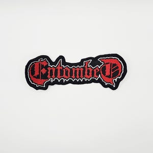 May include: Embroidered patch with the word "Entombed" in red gothic lettering. The patch has a black border with a white outline, set against a white background. The patch is likely for clothing or accessories.