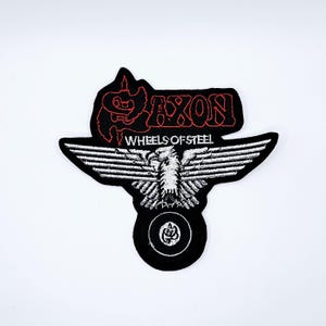 May include: Black and white embroidered patch featuring the word "SAXON" in red, above the words "WHEELS OF STEEL". The patch has a stylized eagle with outstretched wings and a wheel design.