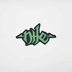 Könnte beinhalten: Gestickter Aufnäher mit dem Wort "DHE" in einer stilisierten gotischen Schrift. Die grünen Buchstaben haben einen schwarzen Rand und sind auf weißem Hintergrund platziert. Der Aufnäher hat ein einzigartiges, künstlerisches Design.