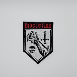 Puede incluir: Un parche en blanco y negro con la palabra "DIOCLETIAN" en rojo. El parche presenta un brazo esquelético sosteniendo una figura demoníaca, una calavera y una cruz. El parche tiene un borde negro y un fondo blanco.