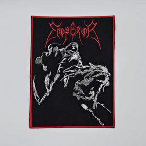 Może przedstawiać: Czarna, haftowana naszywka z czerwoną obwódką. Zawiera nazwę zespołu "EMPEROR" w czerwonych gotyckich literach. Widnieje również biała ilustracja postaci na koniu, częsty motyw w merchandisingu zespołów metalowych.