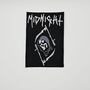 May include: A rectangular black embroidered patch featuring the word MIDNIGHT in white stylized lettering at the top. Below the text is a white outlined diamond shape containing a grey hooded skull figure with a scythe, all on a black background.