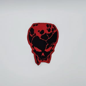 May include: A red and black skull patch. The skull design features a cracked pattern on the top half and glowing red eyes. The patch is likely made of fabric and is designed to be sewn or ironed onto clothing or accessories.