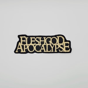 Può includere: Una toppa nera con testo ricamato in oro che recita "FLESHGOD APOCALYPSE". La toppa ha un bordo nero e una forma rettangolare. Il testo è in un carattere in grassetto e stilizzato.