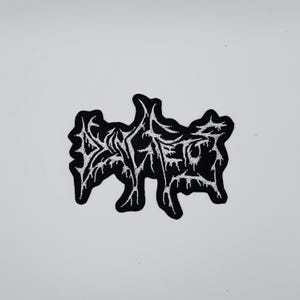 Op de afbeelding: Zwart en wit geborduurde patch met een gestileerd ontwerp. De patch heeft een zwarte achtergrond met witte, druipende letters. De patch is waarschijnlijk voor kleding of accessoires.