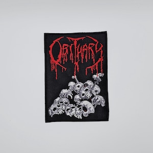 May include: Black rectangular patch featuring the word "OBITUARY" in dripping red letters above a pile of gray skulls. The patch is a heavy metal band merchandise item.