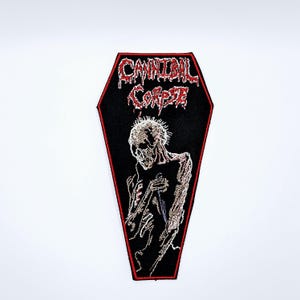 Könnte beinhalten: Schwarzer Sargförmiger Aufnäher mit dem Text "CANNIBAL CORPSE" in roten, tropfenden Buchstaben. Der Aufnäher zeigt ein detailliertes gesticktes Bild einer skelettartigen Figur, die ein Messer hält. Der Rand ist rot.
