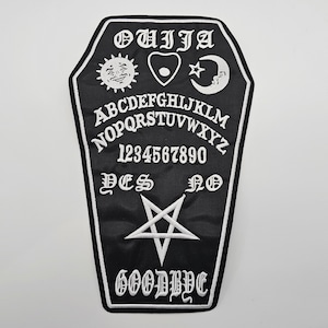May include: Black and white embroidered patch shaped like a coffin with the word "OUIJA" at the top and a Ouija board design with letters, numbers, "YES", "NO", and "GOODBYE" in white.