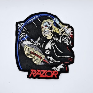 Op de afbeelding: Geborduurde patch met een skeletfiguur op een motorfiets, met het woord "RAZOR" in het rood. De patch heeft een zwarte achtergrond met blauwe en witte accenten en een gouden en rood embleem.