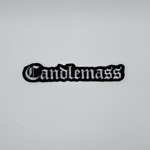 Op de afbeelding: Een zwart-wit geborduurde patch met het woord "Candlemass" in een gotisch lettertype. De patch heeft een zwarte rand en een rechthoekige vorm. De tekst is wit.