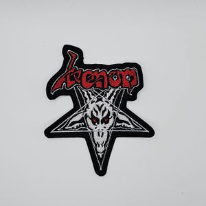 Op de afbeelding: Geborduurde patch met het woord "Venom" in rode, gestileerde letters boven een witte pentagram met een geitenkop en rode ogen. De patch heeft een zwarte rand, voor kleding of accessoires.