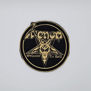 Op de afbeelding: Een ronde, zwarte patch met gouden borduursel. Het woord "Venom" staat in een boog boven een gouden pentagram met een geitenkop. De tekst "Welcome To Hell" staat eronder geborduurd.