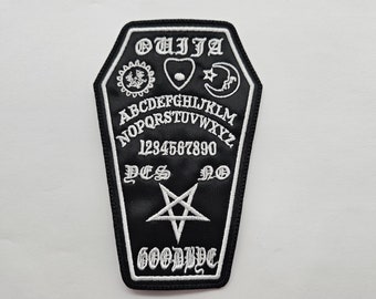 May include: Black and white embroidered patch shaped like a coffin with the word "OUIJA" at the top and a Ouija board design with the letters, numbers, "YES", "NO", and "GOODBYE" inside. A pentagram is in the center of the board.