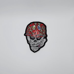 May include: Embroidered patch featuring a skull wearing a red and green camouflage helmet. The helmet has several patches with various symbols. The skull is white with black detailing, and the patch has a black border.