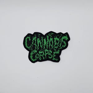May include: A black patch with the words "CANNABIS CORPSE" embroidered in green, stylized with a dripping, organic font. The patch has a black border and is set against a white background.